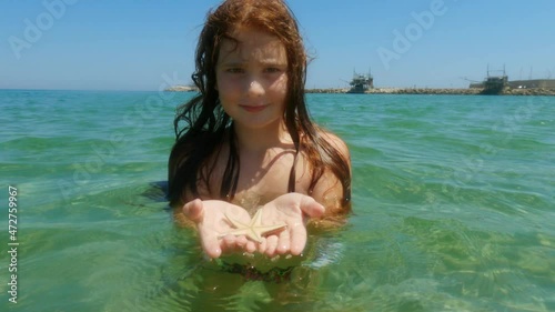 Beautiful adorable redhead child girl bathing in seawater in Italy while holding starfish in hands with trabocchi in background. Slow-motion photo