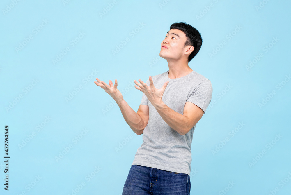 Asian man raising open hands with shocked face and looking at empty space above isolated on studio light blue background