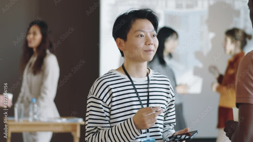 Young Asian woman holding smartphone and glasses while speaking with male colleague on business event Young Asian woman holding smartphone and glasses while speaking with male colleague on business event