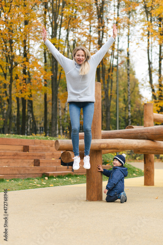 Young mother jumping from the wooden logs on playground while her son is watching in park