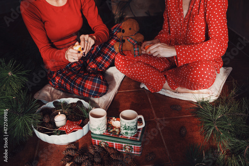 Two young women in red pajamas eating mandarines in cottage, december morning, christmass decoration