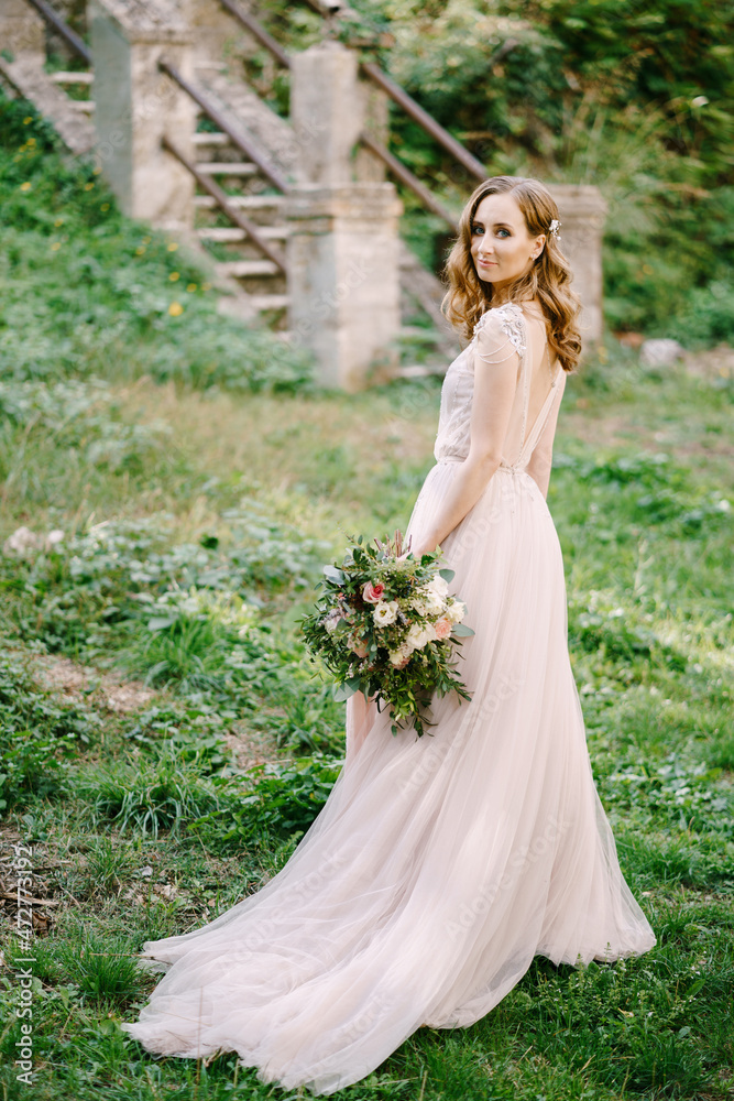 Bride with a bouquet stands half-turned on the green grass in front of a stone staircase