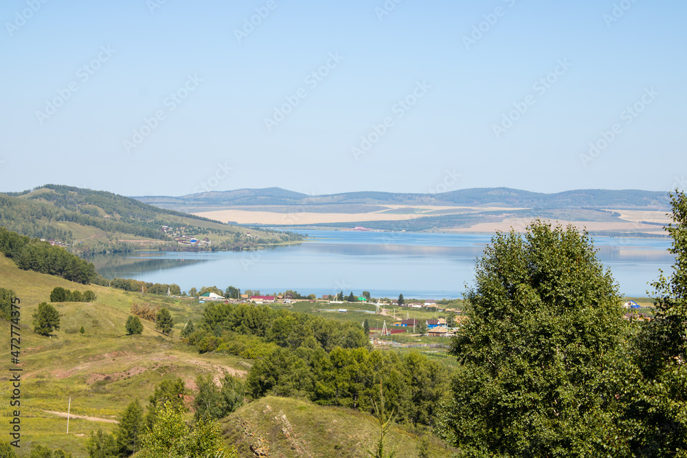 Picturesque landscape of azure lake among the mountains in summer. Village houses next to the lake. Lake Bolshoye, Krasnoyarsk Territory, Siberia, Russia.