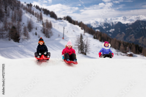 childhood, sledging and season concept - group of happy little kids sliding on sleds down snow hill in winter over alps mountains on background