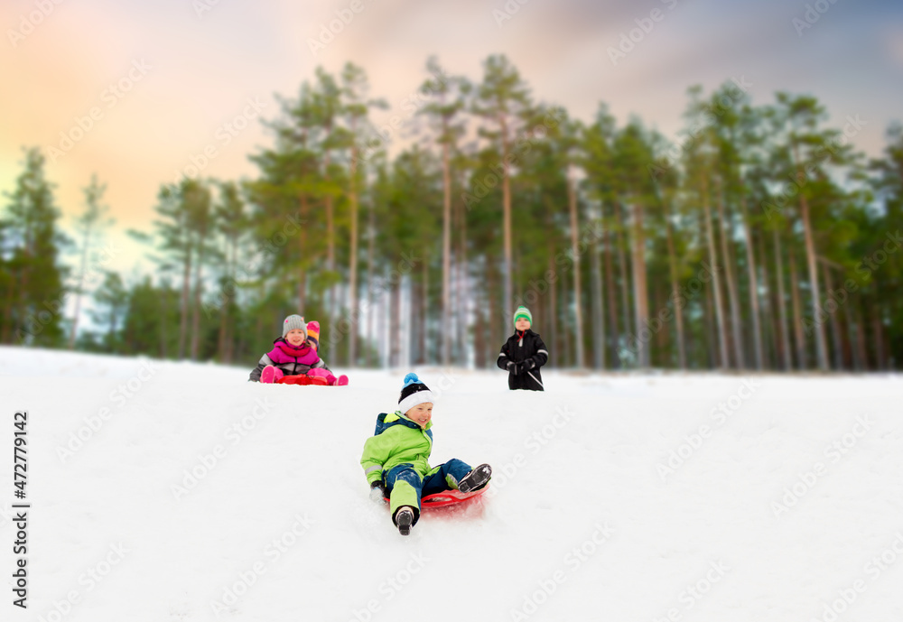 childhood, sledging and season concept - group of happy little children sliding on sleds down snow hill in winter over snowy forest or park background childhood, sledging and season concept - group of happy little children sliding on sleds down snow hill in winter over snowy forest or park background