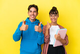 Young student couple isolated on yellow background giving a thumbs up gesture with both hands and smiling