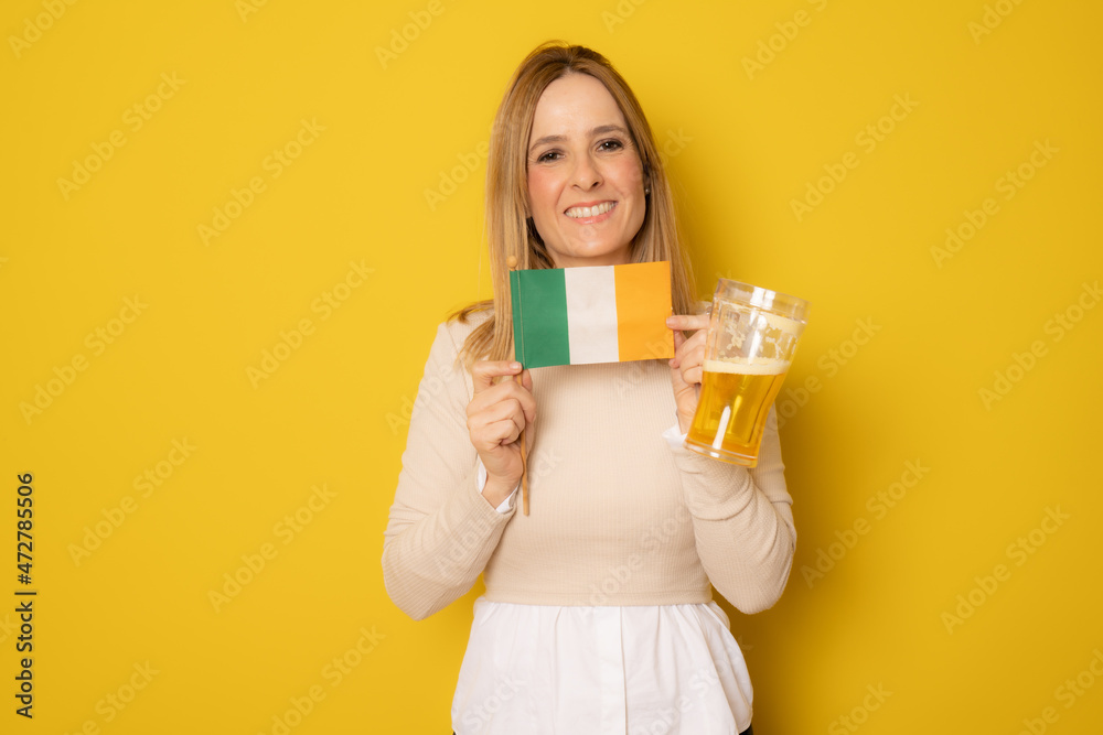 Young smiling woman holding a beer and irish flag standing isolated over yellow background. Young smiling woman holding a beer and irish flag standing isolated over yellow background.