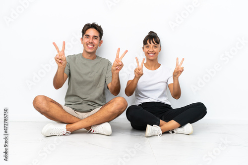 Young mixed race couple sitting on the floor isolated on white background smiling and showing victory sign with both hands