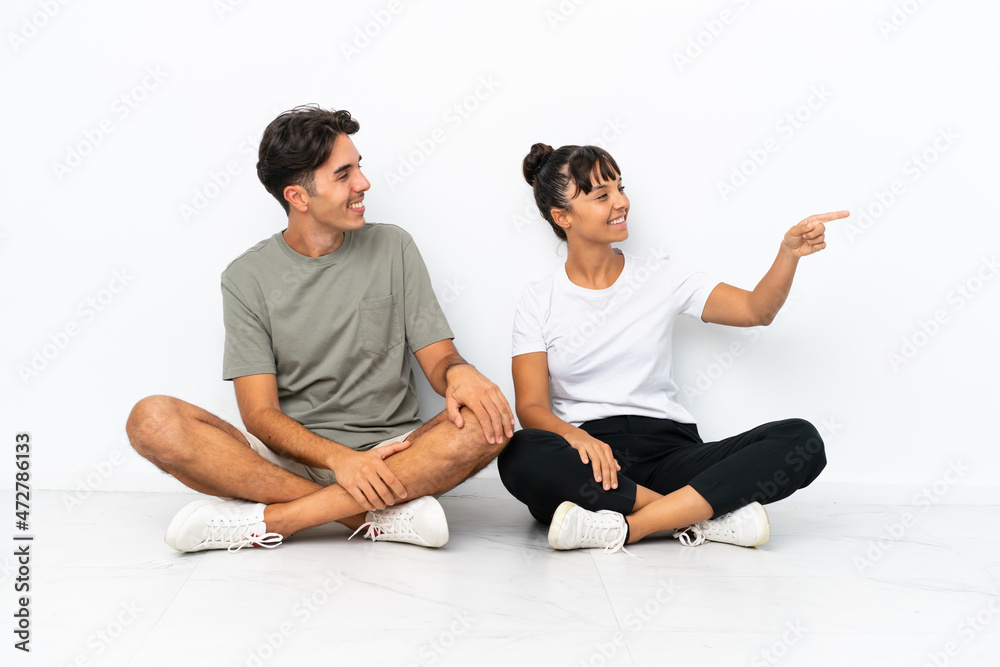 Young mixed race couple sitting on the floor isolated on white background presenting an idea while looking smiling towards Young mixed race couple sitting on the floor isolated on white background presenting an idea while looking smiling towards