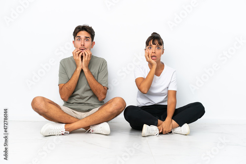 Young mixed race couple sitting on the floor isolated on white background surprised and shocked while looking right