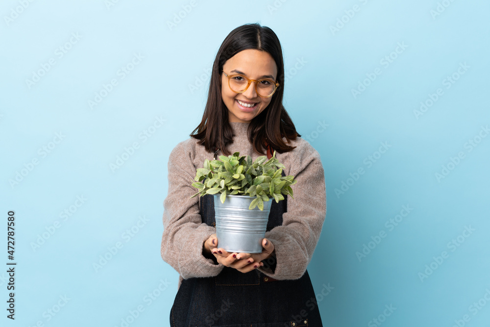 Young brunette mixed race woman holding a plant over isolated blue background holding copyspace imaginary on the palm to insert an ad. Young brunette mixed race woman holding a plant over isolated blue background holding copyspace imaginary on the palm to insert an ad.