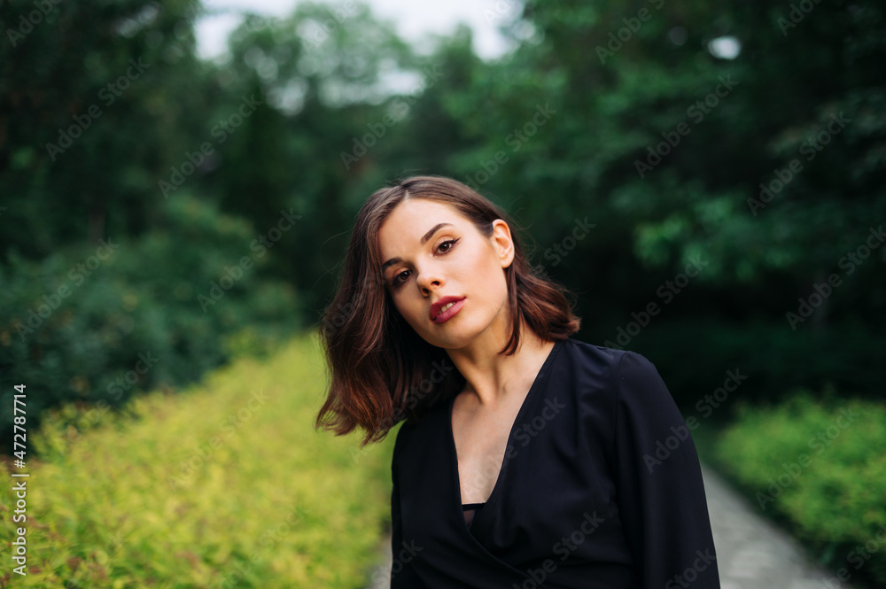 Portrait of a beautiful woman in a black dress with a serious face posing at camera on the background of the evening park.