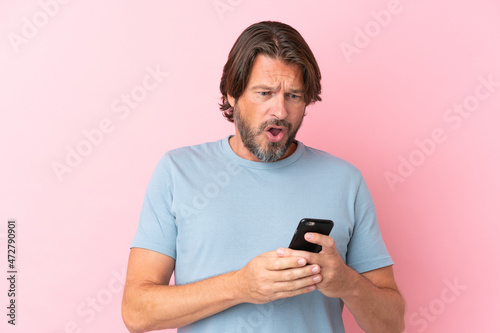 Senior dutch man isolated on pink background looking at the camera while using the mobile with surprised expression © luismolinero