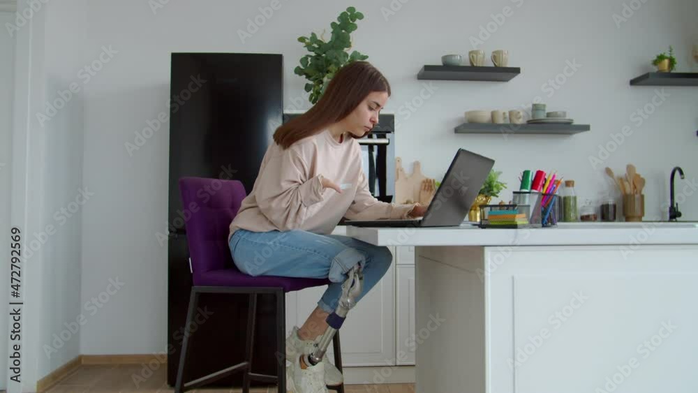 Charming positive young woman with prosthetic leg and hand birth anomaly networking on laptop, browsing social media content online while spending leisure in domestic kitchen. Charming positive young woman with prosthetic leg and hand birth anomaly networking on laptop, browsing social media content online while spending leisure in domestic kitchen.