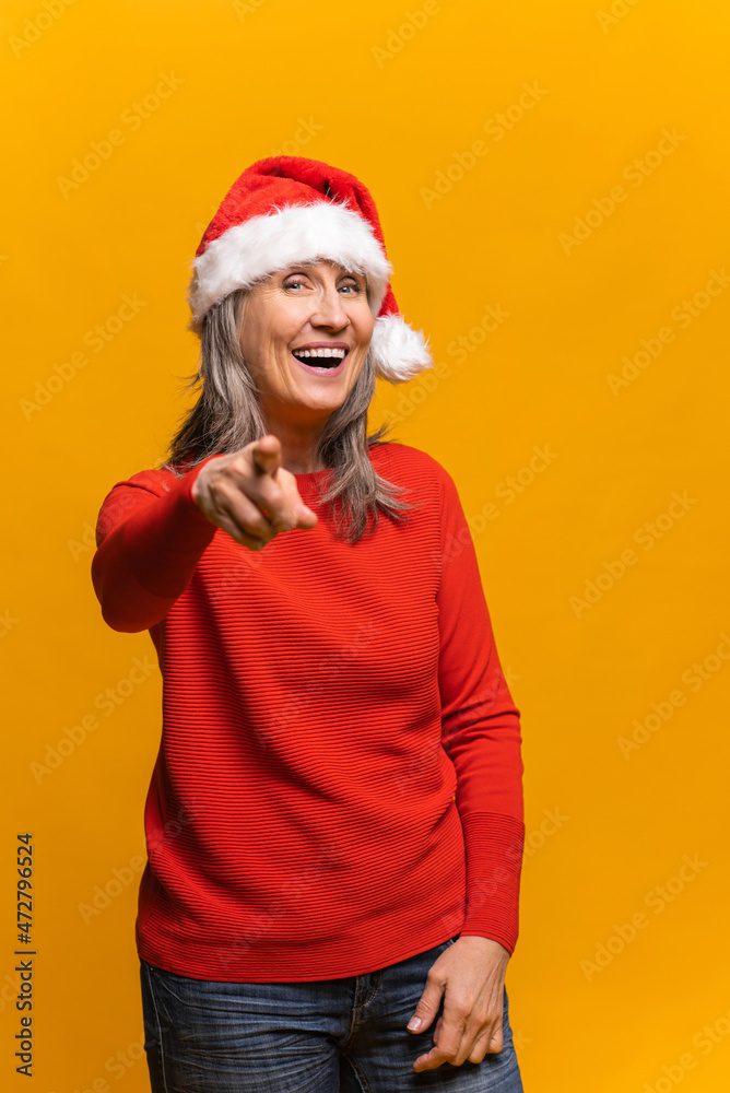 Cheerful happy senior woman in santa hat points index finger at camera and laughs, vertical portrait middle-aged female choosing you. New year advertising concept Cheerful happy senior woman in santa hat points index finger at camera and laughs, vertical portrait middle-aged female choosing you. New year advertising concept