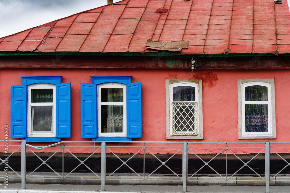 An old wooden house with blue shutters and platbands and an iron roof. The picture was taken in Russia, in the city of Orenburg