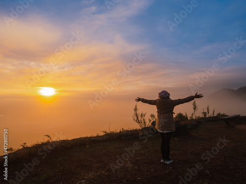 back of Asian woman with freedom arms open standing on mountain peak at Pha Tang view point 104 with river view and sea mist and sun rise in fog sky. travel place in Chaing Rai, Thailand