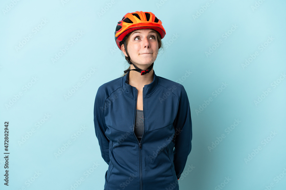 Young cyclist woman isolated on blue background and looking up - obrazy, fototapety, plakaty 