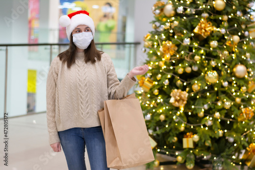 Woman wearing a facemask in a santa claus hat, sweater in a shopping center near the christmas tree. Concept: holiday shopping during the coronavirus epidemic a woman wearing a medical mask 