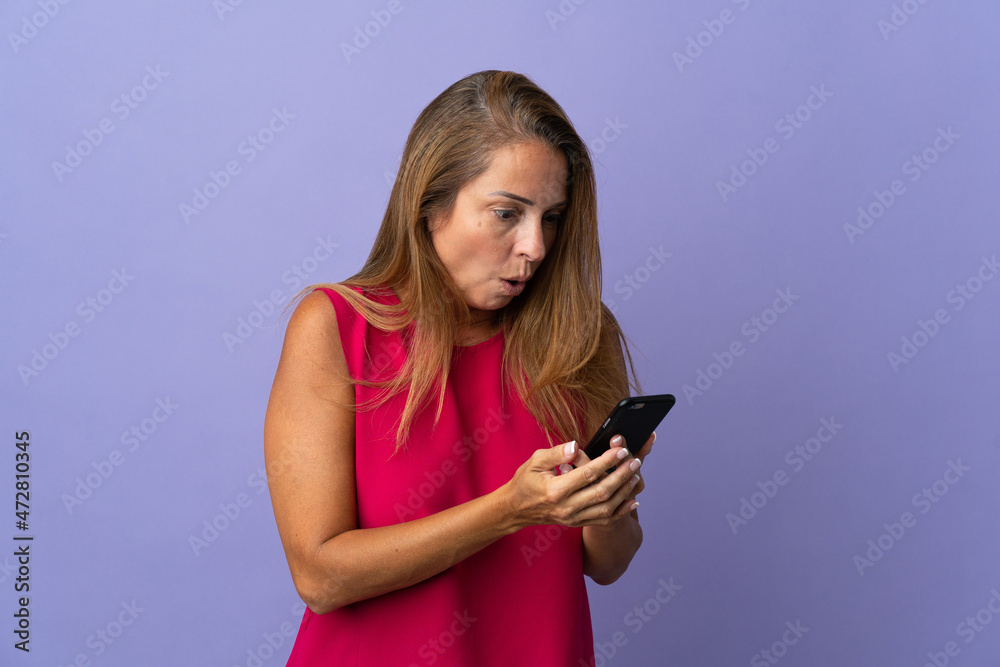 Middle age brazilian woman isolated on purple background looking at the camera while using the mobile with surprised expression Middle age brazilian woman isolated on purple background looking at the camera while using the mobile with surprised expression