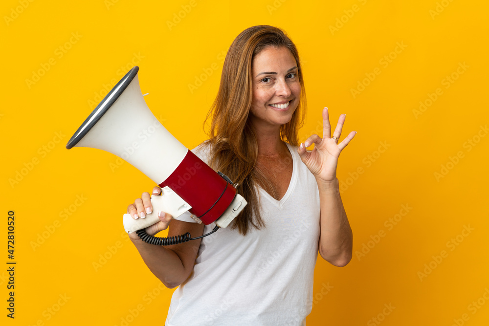 Middle age brazilian woman isolated on yellow background holding a megaphone and showing ok sign with fingers Middle age brazilian woman isolated on yellow background holding a megaphone and showing ok sign with fingers