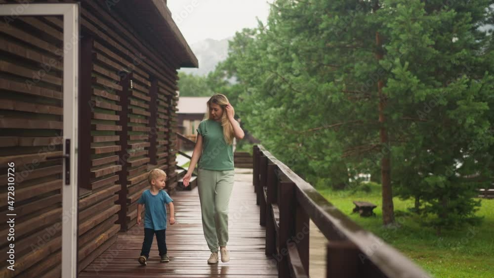Blonde mother gives hand to offended weepy little boy walking along veranda deck to hotel room at resort on rainy summer day slow motion Blonde mother gives hand to offended weepy little boy walking along veranda deck to hotel room at resort on rainy summer day slow motion