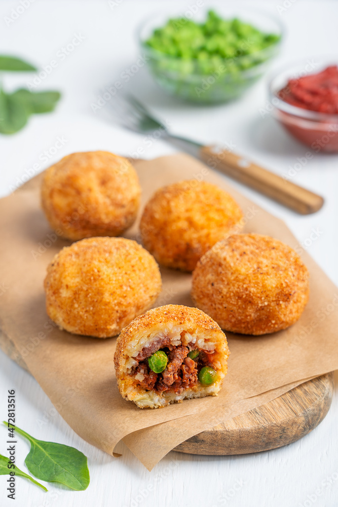 Homemade Arancini Italian rice balls deep fired and stuffed with minced beef meat in tomato sauce and green peas served on brown wooden board with fork and spinach leaves on white table. Vertical