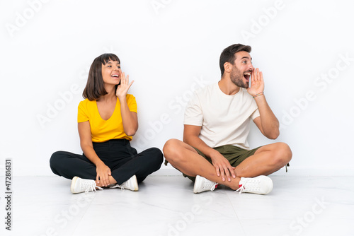Young couple sitting on the floor isolated on white background shouting with mouth wide open to the lateral