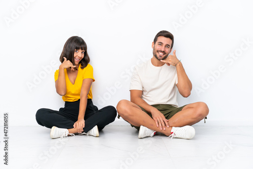 Young couple sitting on the floor isolated on white background making phone gesture. Call me back sign