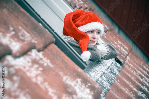 Bilde på lerret Young woman in a red and white Christmas hat looking out of a skylight window waiting for a miracle or just for a first snowfall