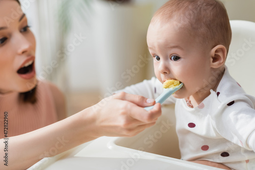 toddler kid in baby chair eating puree near blurred mother.