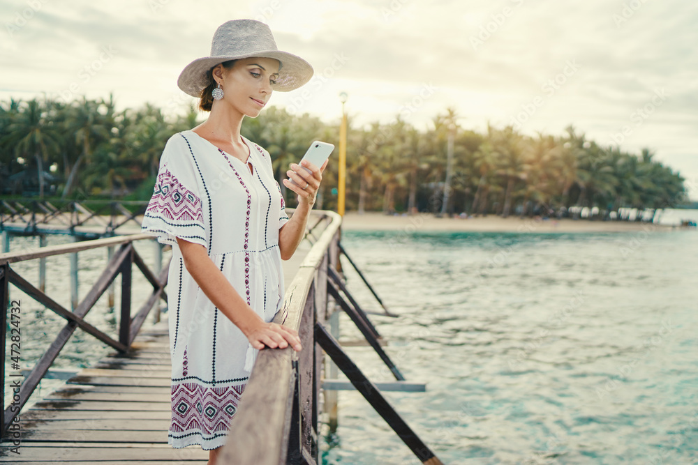Vacation and technology. Young pretty woman in hat using smartphone while walking on wooden brige with tropical island view.