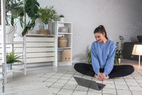 Happy attractive young woman in active wear sitting on mat in lotus position, starting online training video call yoga class with trainer or performing webinar session as coach distantly. Home workout