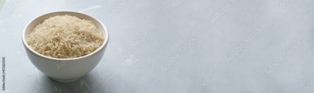 Baner. White milled long grain rice in a white bowl on a gray background. Vegan food concept. Rice dishes are cooked in all countries of the world. Popular cereal. Copy space. Baner. White milled long grain rice in a white bowl on a gray background. Vegan food concept. Rice dishes are cooked in all countries of the world. Popular cereal. Copy space.