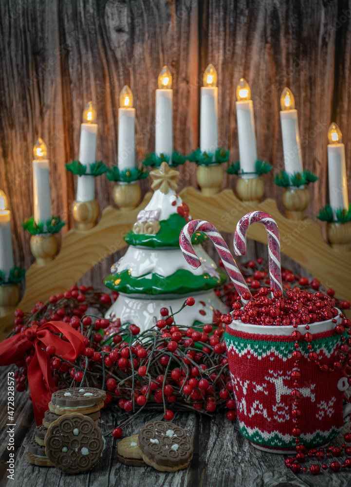 Fototapeta premium Moscow, Russia, December 2021, Christmas card, background, cup in a sweater with beads and candy canes on the background of a Christmas wreath of red berries, a ceramic tree and a candlestick