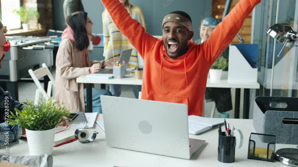 Joyful Afro-American businessman is sharing good news with colleagues laughing doing high-five expressing emotions in modern coworking space. Joyful Afro-American businessman is sharing good news with colleagues laughing doing high-five expressing emotions in modern coworking space.