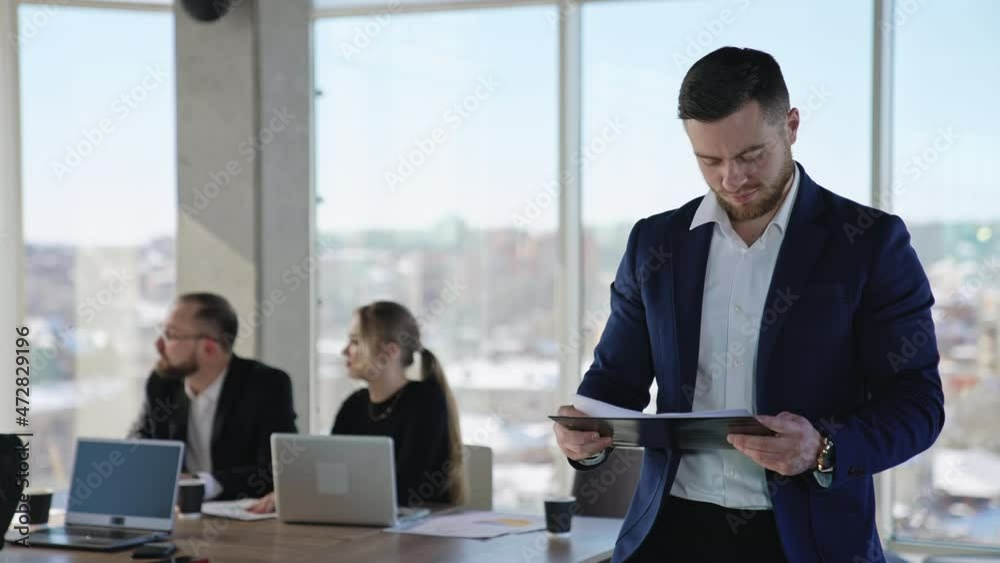 Young businessman in a suit holding the papers and looking at them. Young entrepreneur among the business partners at work. Team of coworkers at the background. Young businessman in a suit holding the papers and looking at them. Young entrepreneur among the business partners at work. Team of coworkers at the background.