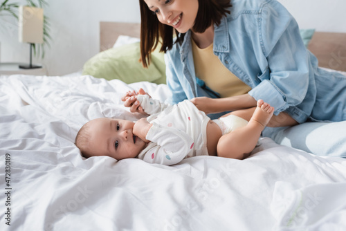 toddler kid holding hand in mouth while lying on bed near cheerful mother.