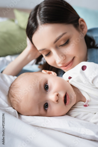 close up view of toddler boy looking at camera while lying near blurred happy mom.