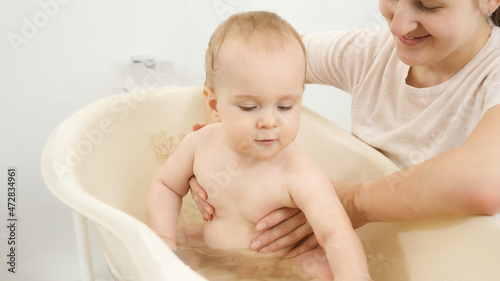 Young mother washing her baby son in small child bath at home