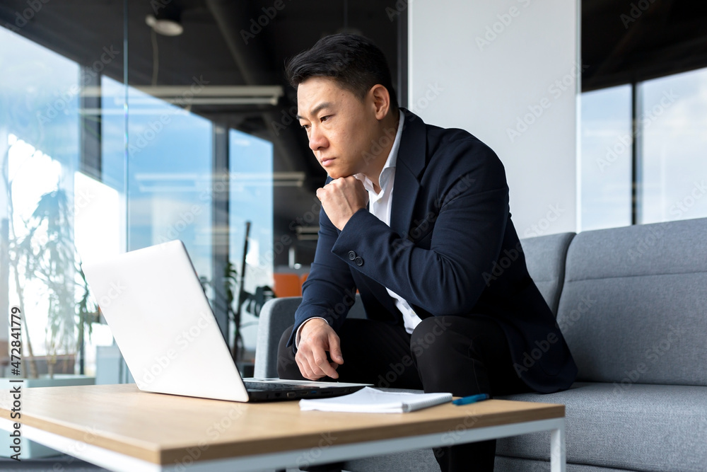 Asian pensive businessman at laptop, thinking about creating a new business strategy, successful man working on laptop at office