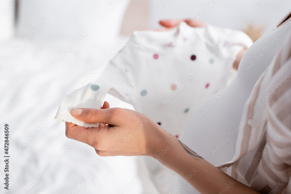cropped view of tattooed pregnant woman holding baby romper in bedroom. cropped view of tattooed pregnant woman holding baby romper in bedroom.