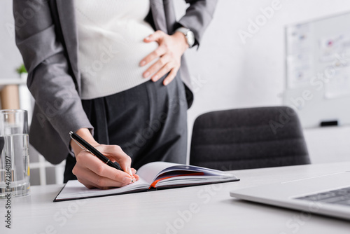 cropped view of pregnant businesswoman writing in notebook.