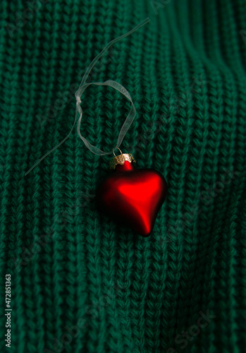 A bright red Christmas tree toy in the shape of a heart on a knitted green sweater.