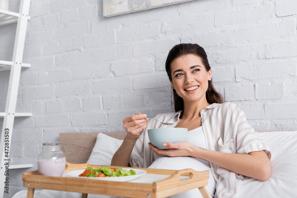 happy pregnant woman holding bowl with oatmeal near tray with salad.