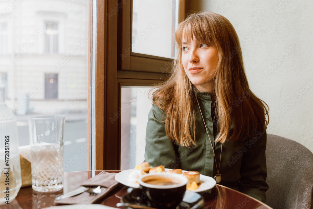 Smiling and thoughtful young female looking away in cafe while drinking coffee and eating cheesecake Smiling and thoughtful young female looking away in cafe while drinking coffee and eating cheesecake