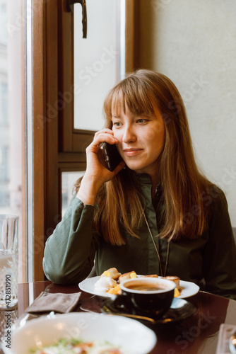 Young female talking on phone while eating dessert and drinking coffee in cafe