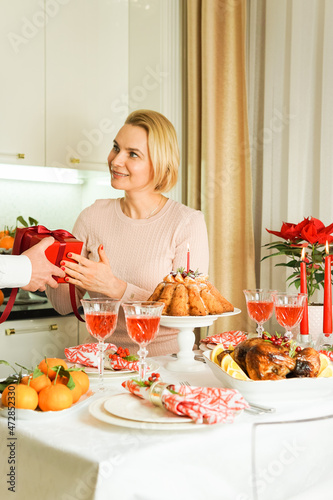 A nice 40-year-old woman is sitting at a festive table, smiling and receiving a gift. Christmas Holiday, Thanksgiving Day, New Year. Festive red gift box.