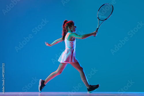 Studio shot of little girl, beginner tennis player training isolated on blue studio background in neon light. Sport, study, childhood concept © master1305