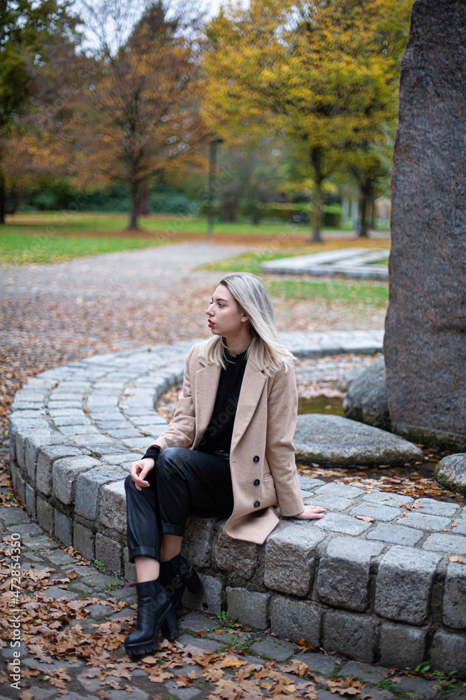 Serious white young european woman with blonde hair in park sitting on stone fence in beige fall coat